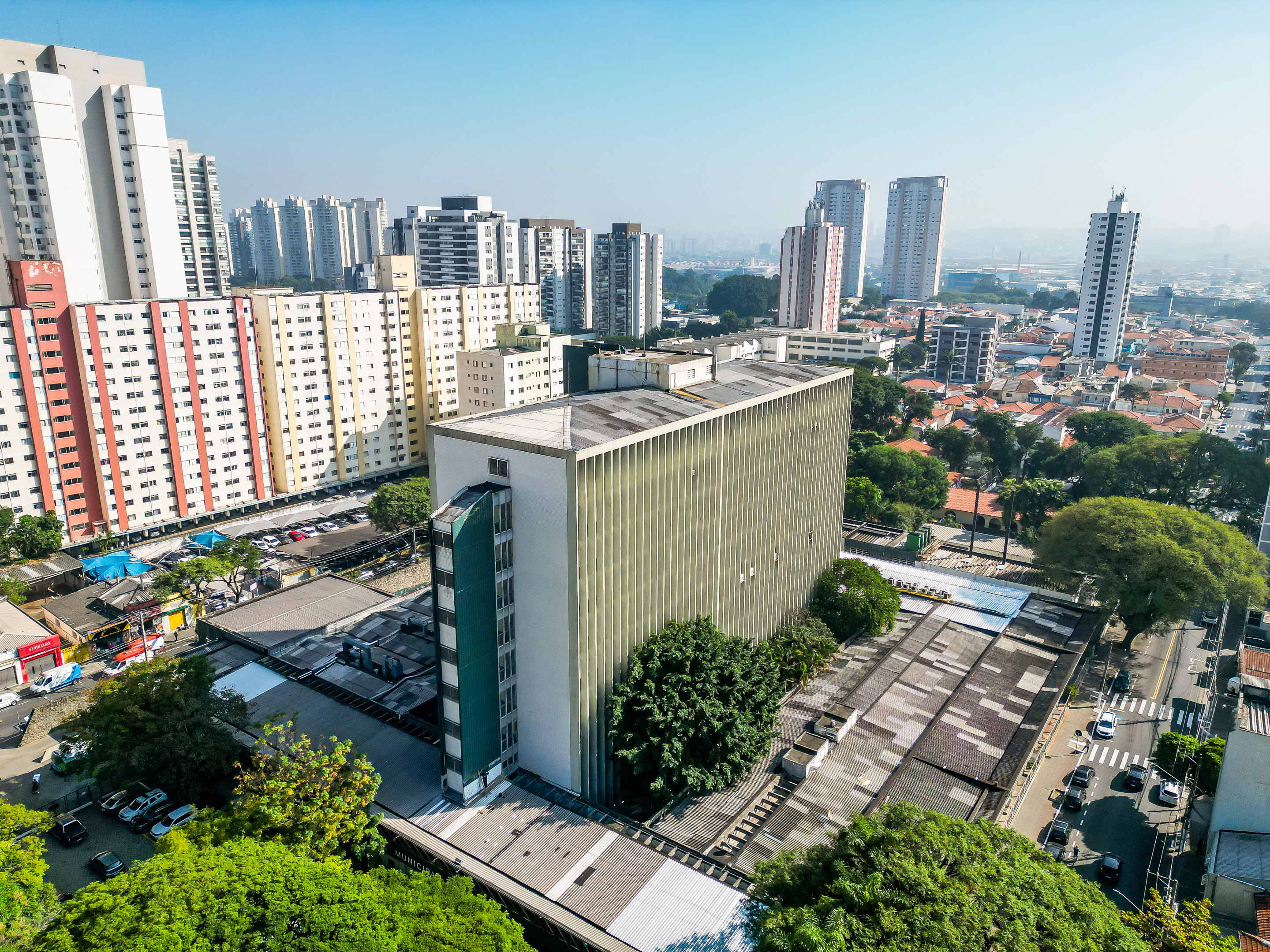 A foto mostra a vista aérea do Hospital Municipal do Tatuaé, um prédio grande, com cerca de sete andares; a visão é panorâmica, e  vemos outros prédios e casas em volta do hospital